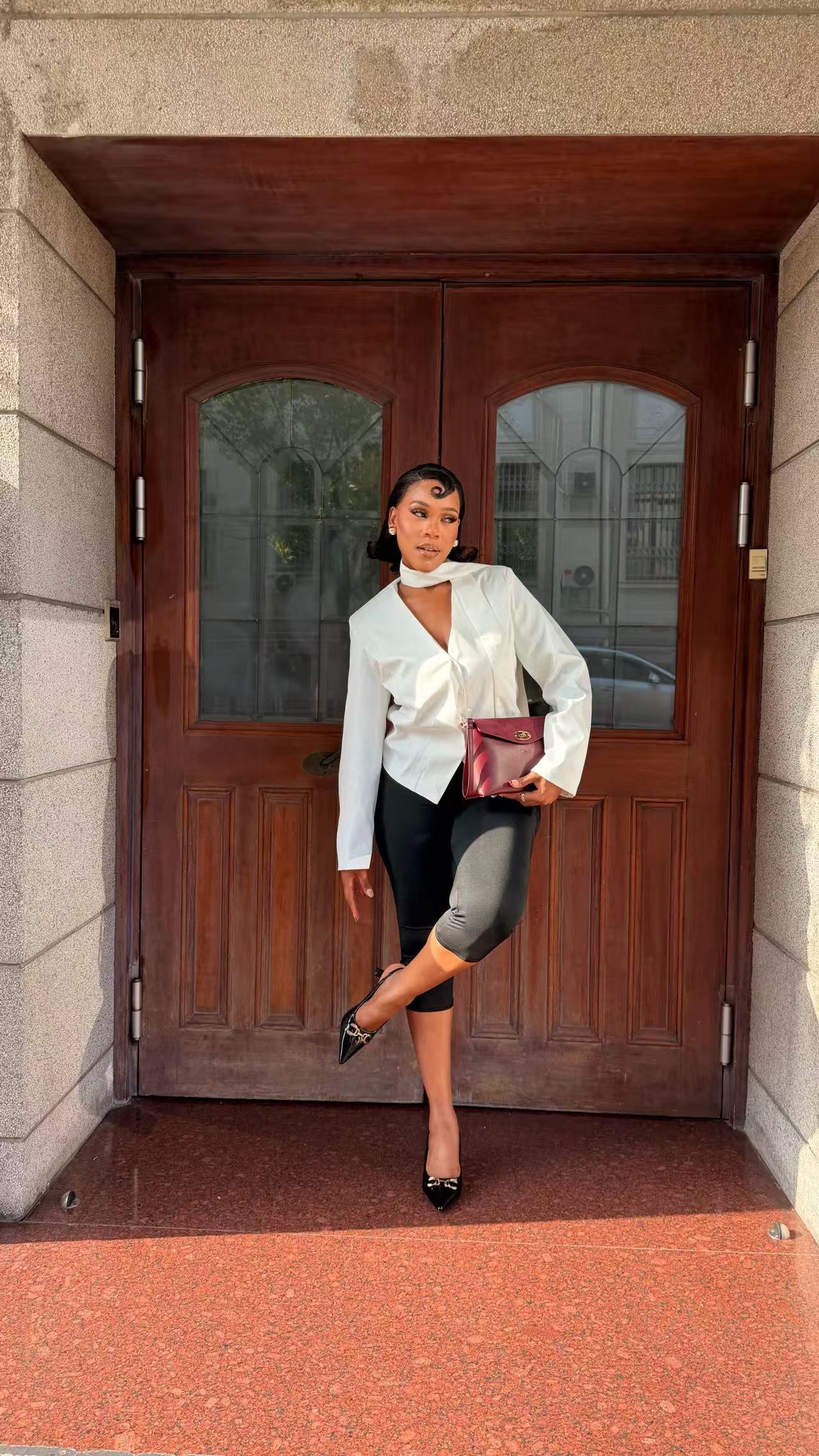 Model in a white blouse carrying the burgundy Adaline bag in a stone doorway